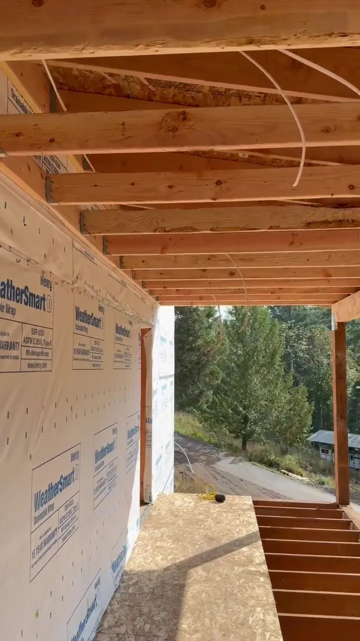 Covered porch of a new ADU near Kalama WA with house wrap on the wall, open ceiling joists with lighting feeds run through them, and a gravel drive and evergreen forest beyond