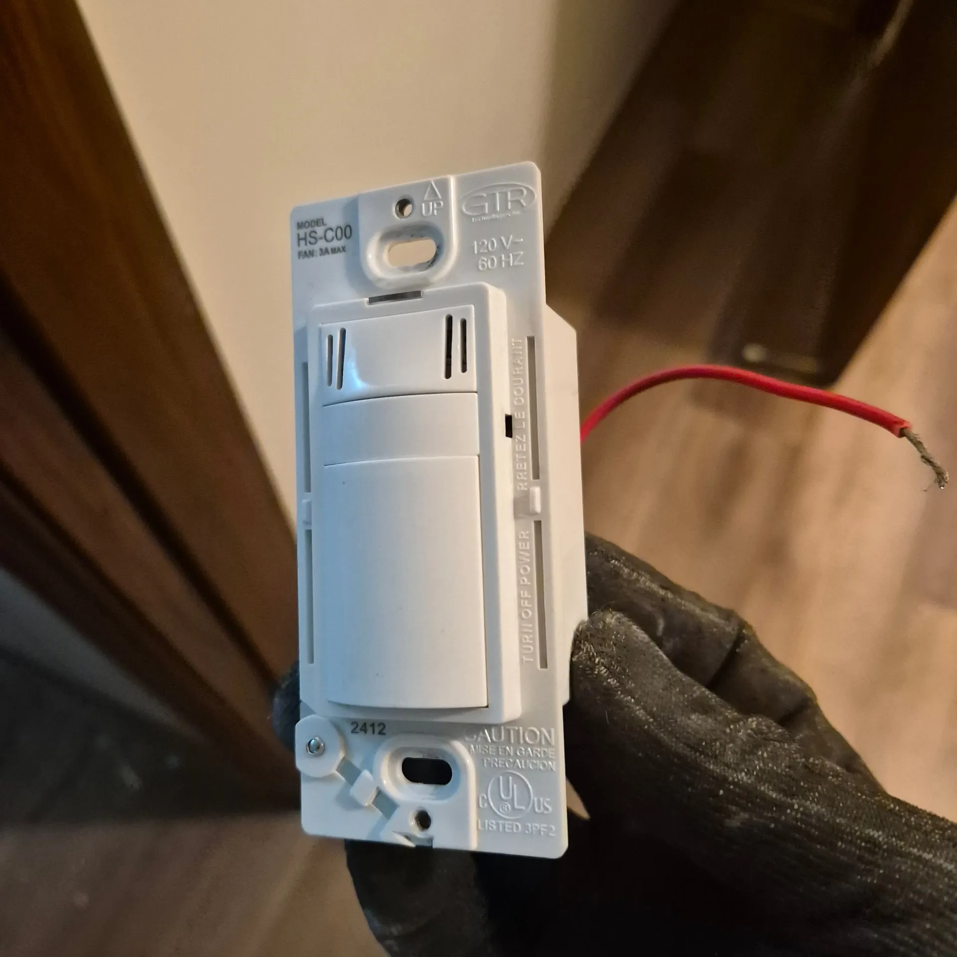 Humidity-sensing bathroom fan switch held in a gloved hand during the repair in a Vancouver WA rental, a red wire connected to the device