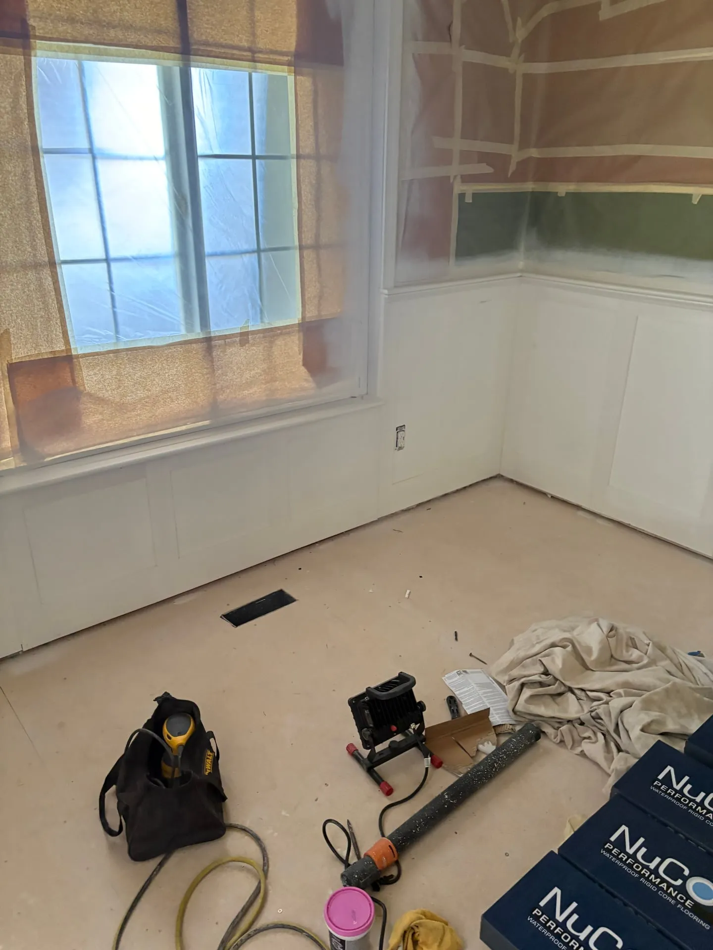 Room in a Vancouver WA home with white board-and-batten wainscoting and masked windows, an open receptacle box at the wall, and boxes of NuCore waterproof flooring stacked ready to install