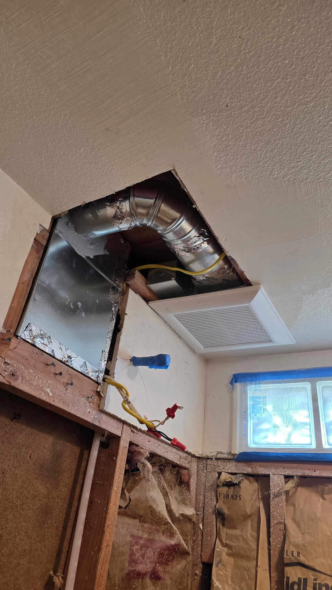 Bath fan installed in the ceiling of a 1920s Hazel Dell bathroom remodel in Vancouver WA, with rigid metal ducting routed through the open wall cavity and a yellow Romex drop to the new fan switch, old newspaper insulation visible in the exposed stud bays
