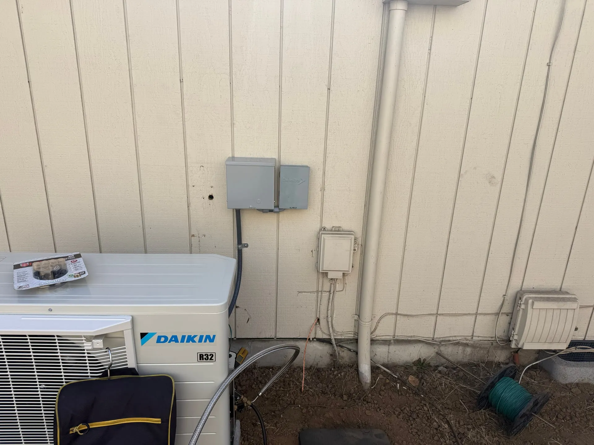 New Daikin R32 heat pump outdoor unit beside the siding of a Vancouver WA home, with a gray outdoor disconnect and a second protective device mounted on the wall above it, flexible conduit running down to the unit