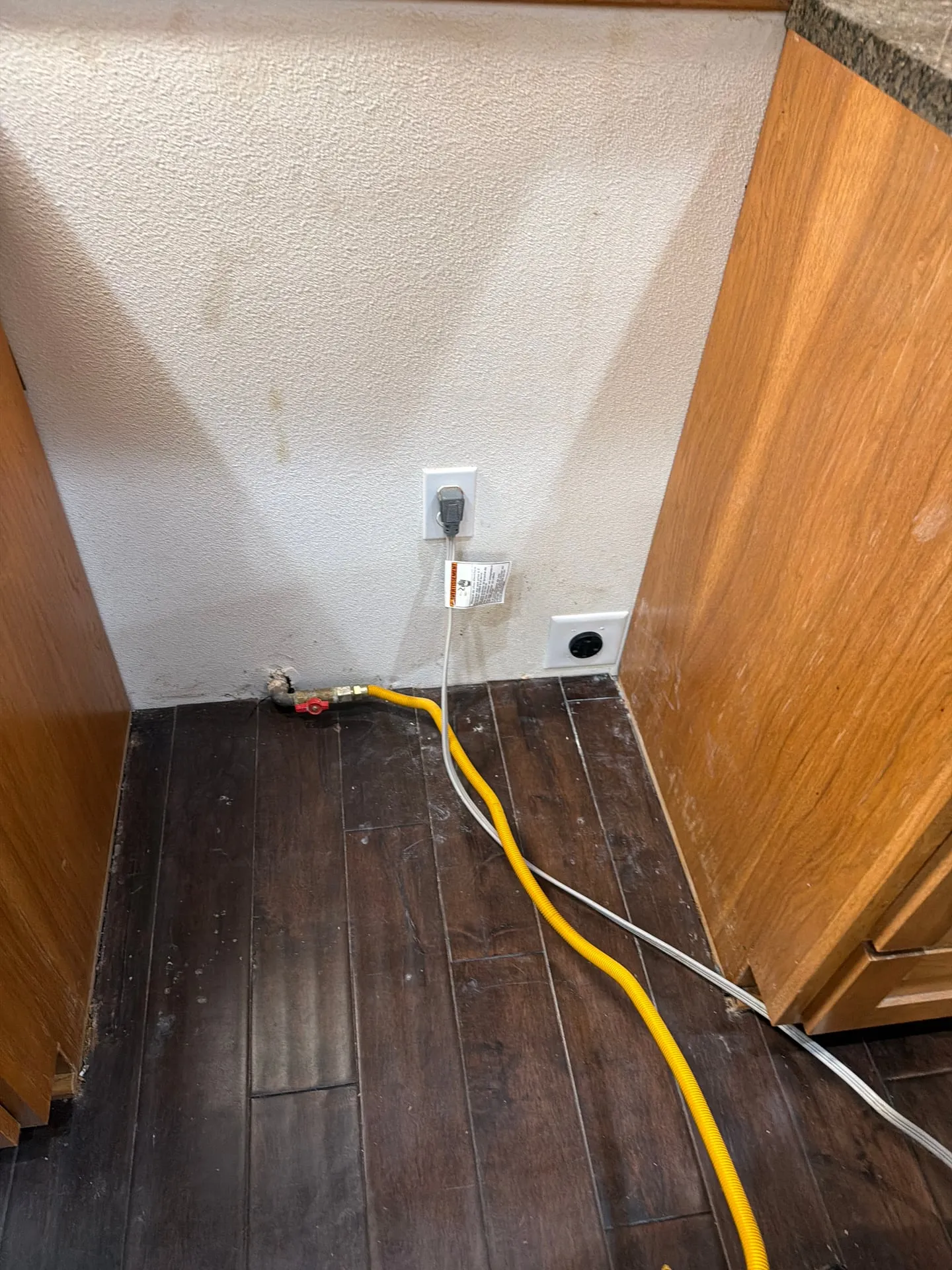 Existing kitchen range location in an Orchards neighborhood home in Vancouver WA before the circuit relocation, showing the old 240V receptacle, a yellow flexible gas supply line with shutoff valve, and a 120V kitchen outlet along the baseboard