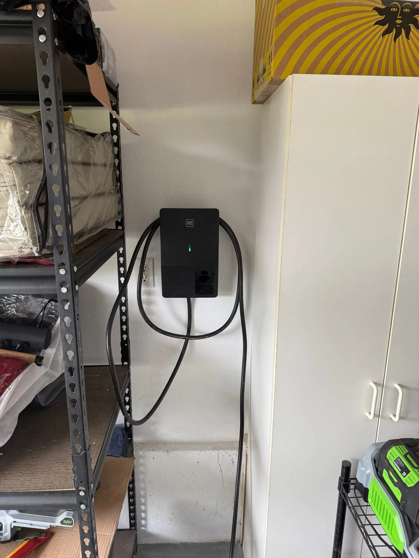 GM-branded Level 2 EV charger mounted on the garage wall of a Camas WA home with green status light on and charging cable coiled, adjacent duplex outlet and garage storage shelving visible