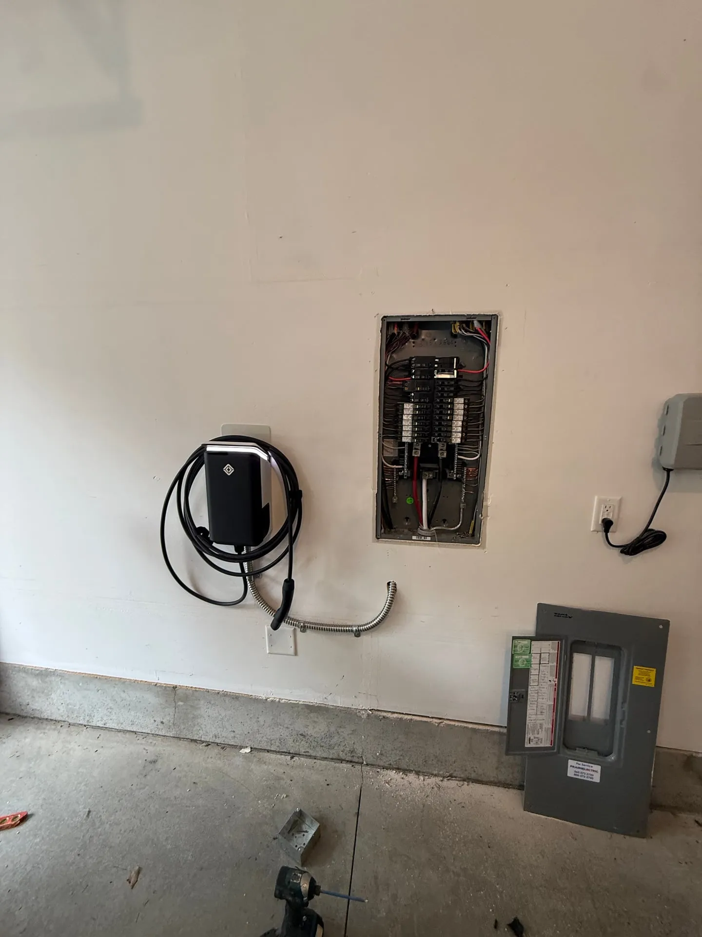 Hardwired black Level 2 EV charger mounted on a garage wall directly beside the open Square D QO electrical panel in a 2022 Vancouver WA home, fed by a short flexible metal whip with the charging cable coiled on its hook