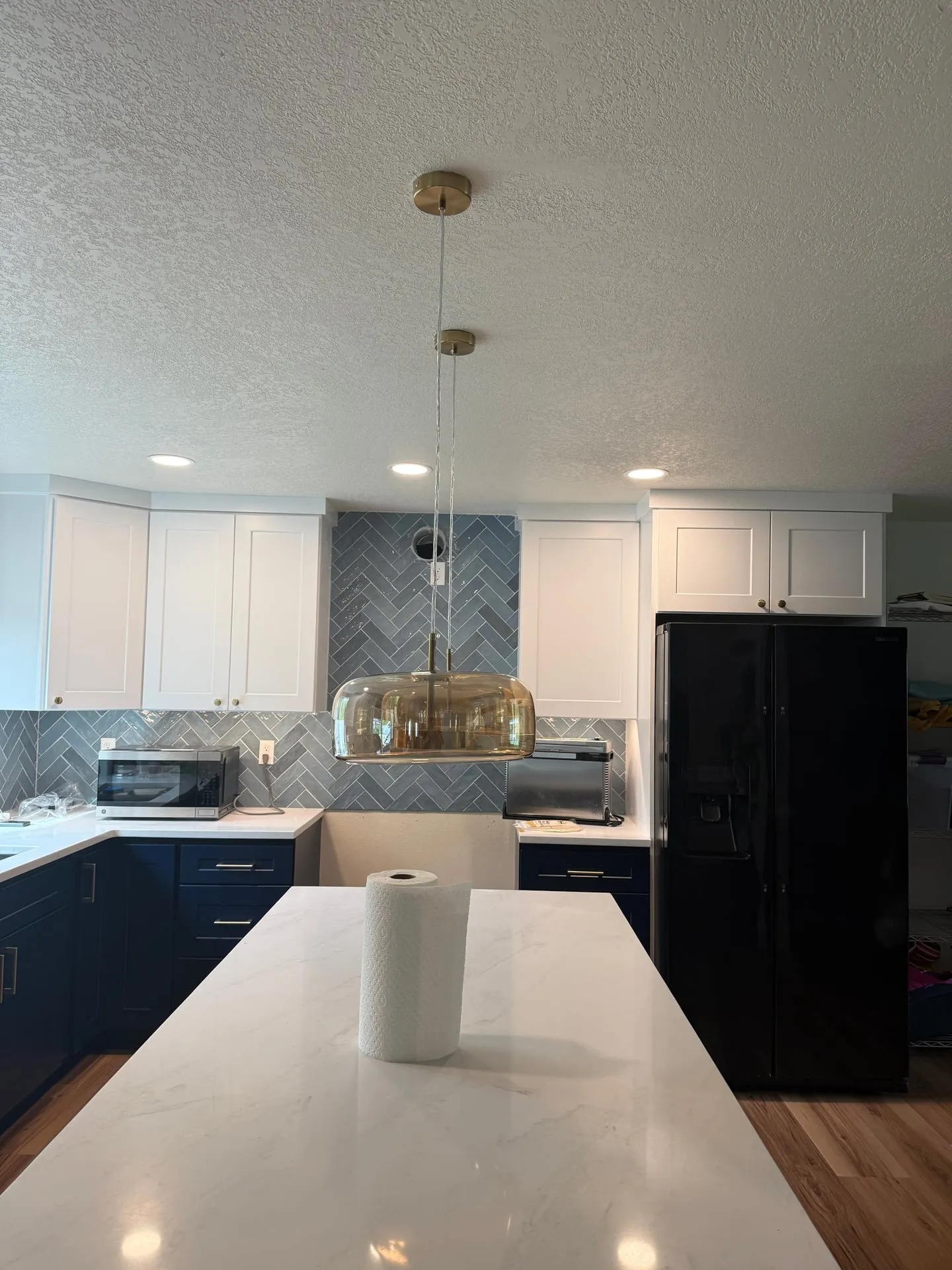 Remodeled kitchen in a Hazel Dell home in Vancouver WA with two amber glass pendants hung over a white quartz island, blue herringbone tile backsplash, white upper cabinets and navy blue lower cabinets, recessed LED cans, and a black Samsung refrigerator