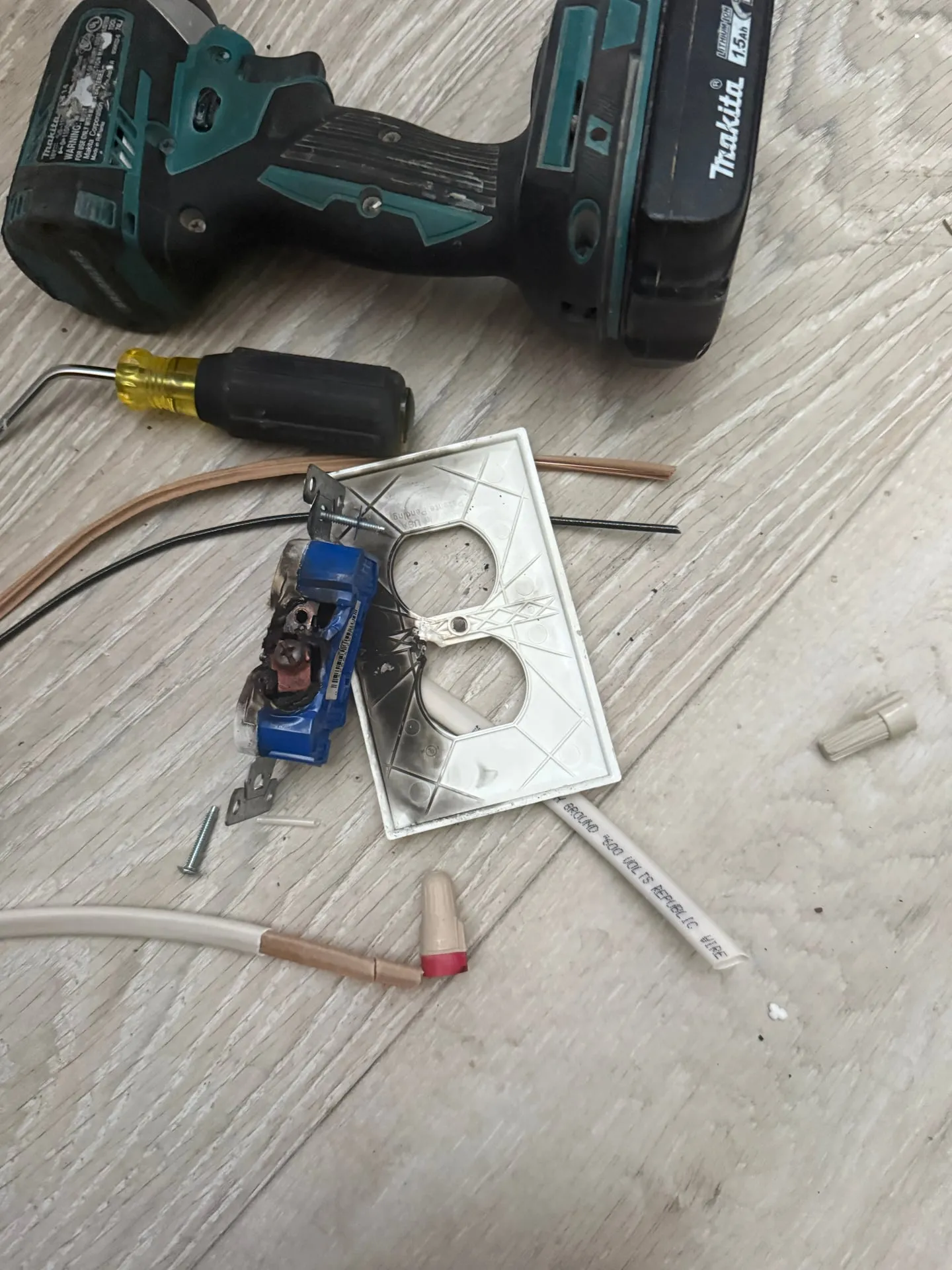 Melted blue electrical receptacle with scorched contacts and a soot-stained cover plate lying on the floor of a Vancouver WA home after being removed from the wall, with a drill and screwdriver beside it