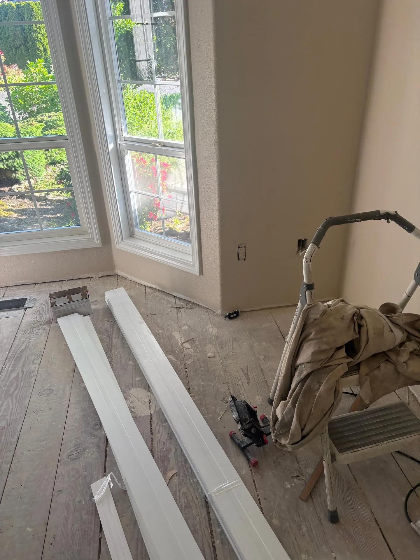 Bay-window room in a Vancouver WA home mid-restoration with the flooring removed to bare subfloor, an open receptacle box at the baseboard waiting for a new outlet, and fresh white trim stock staged on the floor