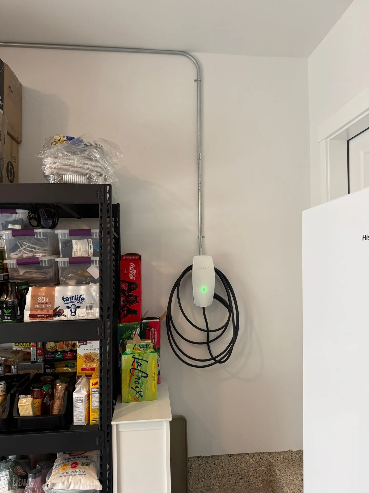 White Tesla Universal Wall Connector mounted on a garage wall in a Ridgefield WA home with a green status light lit, fed by EMT conduit dropping down from the ceiling and the charging cable coiled neatly on its hook