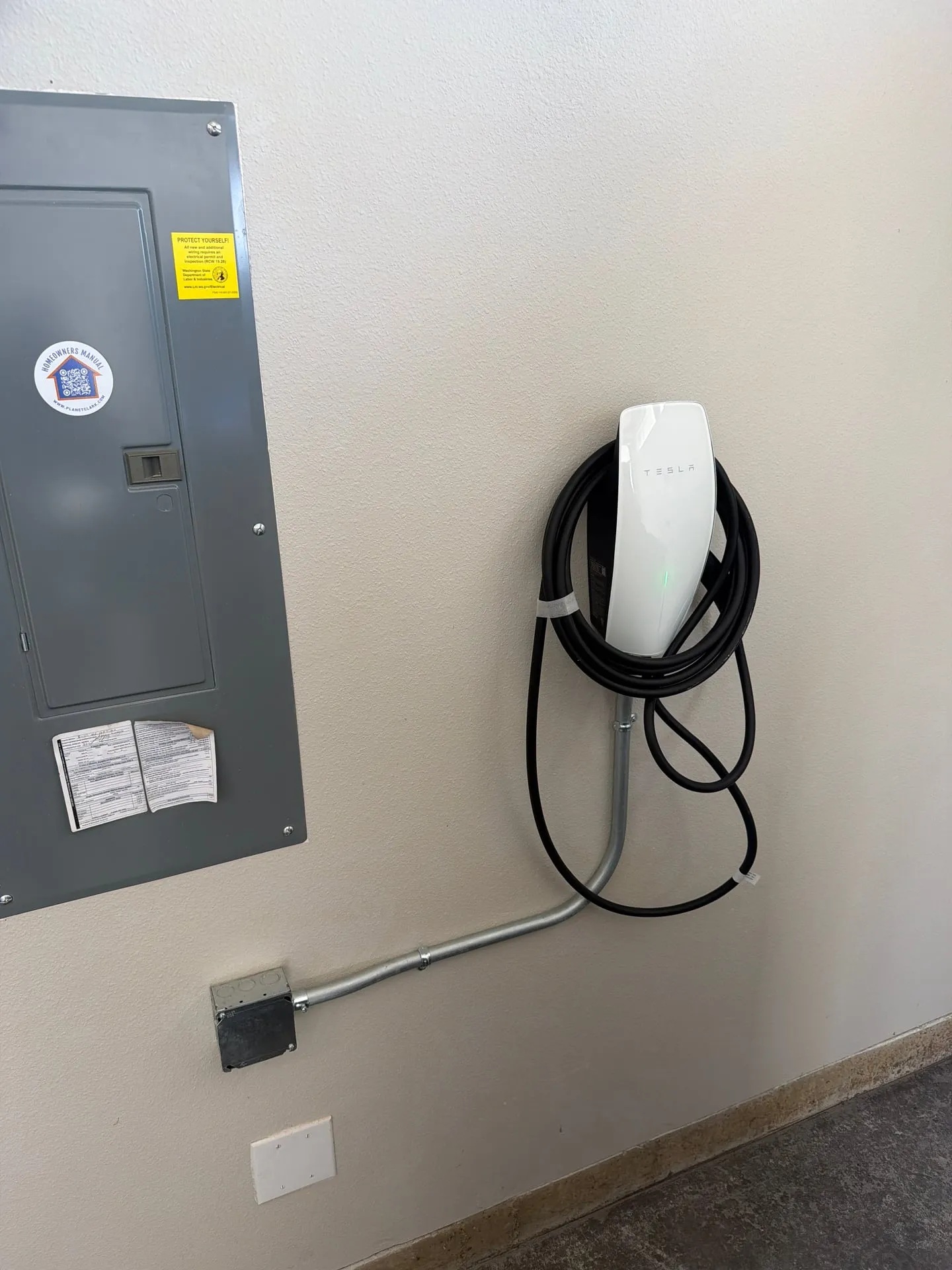 White Tesla Wall Connector mounted on a garage wall next to the electrical panel in a 2022 Vancouver WA home, fed by a short EMT conduit run from the panel up to a junction box and into the charger, charging cable coiled neatly on its hook