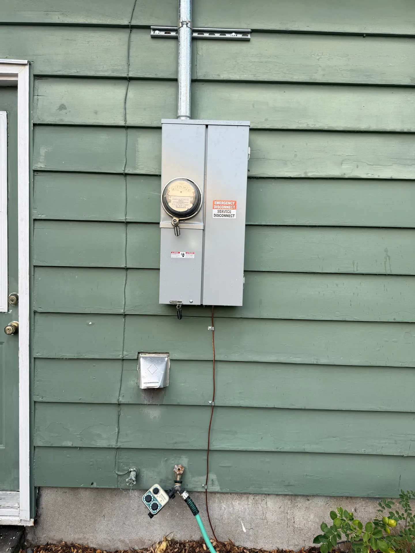 Closed 200-amp outdoor meter main on green siding with an orange Emergency Disconnect and Service Disconnect label on the cover
