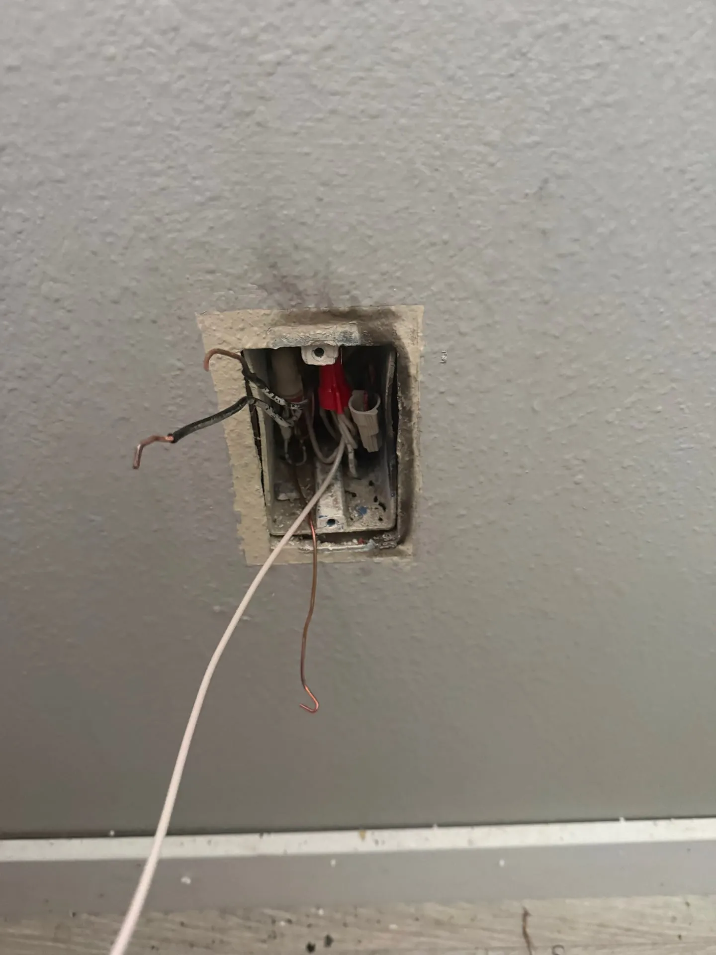 Open electrical box in a gray wall with soot staining around the opening and conductors pulled out after the melted outlet was removed